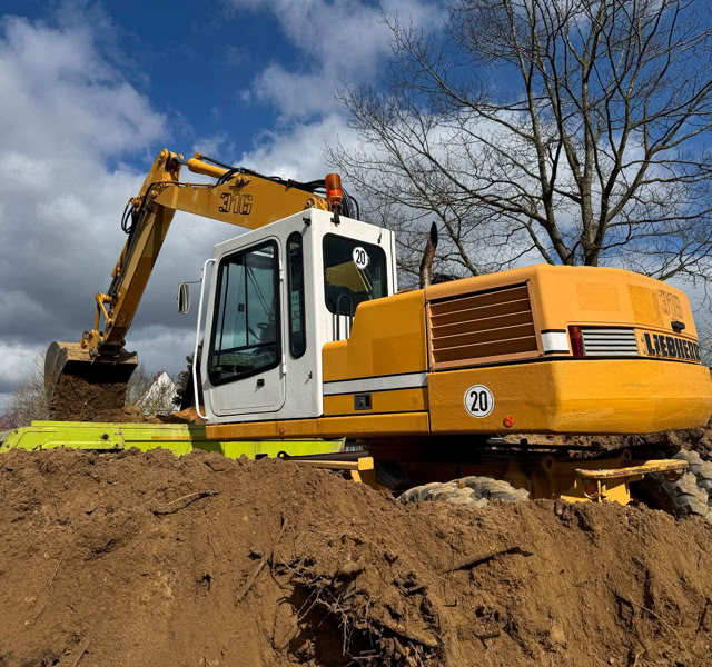 Gelber Bagger auf einer Baustelle, umgeben von Sandhaufen, mit Bäumen und bewölktem Himmel im Hintergrund.