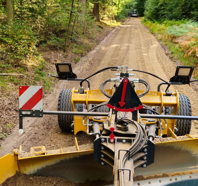 Eine gelbe Baumaschine auf einem unbefestigten Waldweg, umgeben von Bäumen.