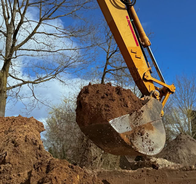 Ein gelber Bagger hebt eine Schaufel voll Erde vor einem blauen Himmel mit einigen Wolken und kahlen Bäumen im Hintergrund.